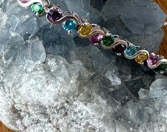 May include: A silver bracelet with a row of colorful gemstones. The gemstones are set in a wavy pattern and include pink, green, blue, purple, and yellow.