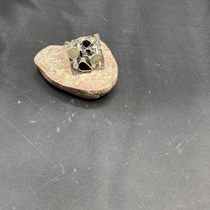 May include: A silver ring with a rectangular design, featuring black, beige, and clear stones in various shapes. The ring is displayed on a small, light brown rock against a dark background. The ring's design includes vertical and horizontal bars.