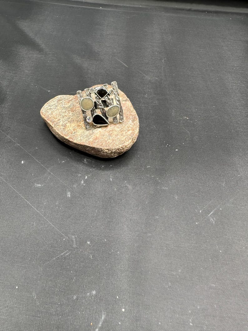 May include: A silver ring with a rectangular design, featuring black, beige, and clear stones in various shapes. The ring is displayed on a small, light brown rock against a dark background. The ring's design includes vertical and horizontal bars.