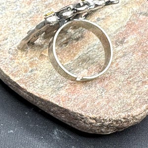 May include: Silver ring featuring a floral design with small yellow gemstones. The ring sits on a textured, light brown stone surface, against a dark background. The ring's band is a simple, polished silver.