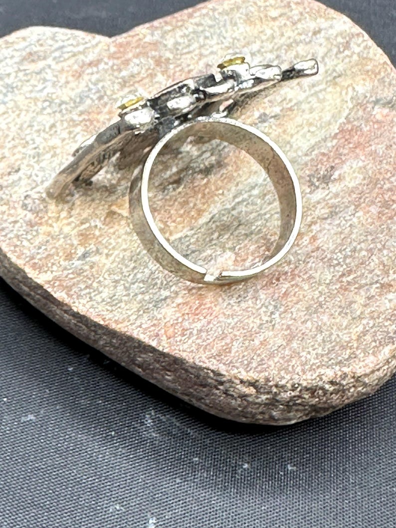 May include: Silver ring featuring a floral design with small yellow gemstones. The ring sits on a textured, light brown stone surface, against a dark background. The ring's band is a simple, polished silver.