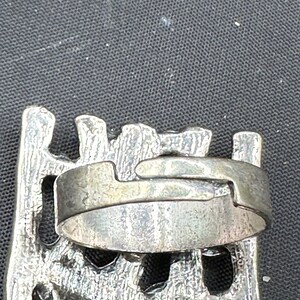 May include: A silver ring with a textured band and an unusual interlocking design. The ring is set against a dark background, highlighting its metallic sheen and intricate details. The ring's design suggests a vintage or artisan style.