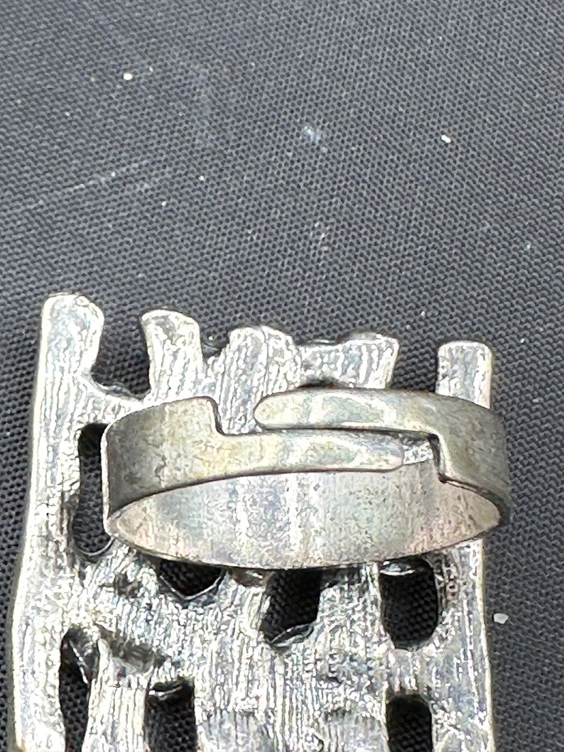 May include: A silver ring with a textured band and an unusual interlocking design. The ring is set against a dark background, highlighting its metallic sheen and intricate details. The ring's design suggests a vintage or artisan style.