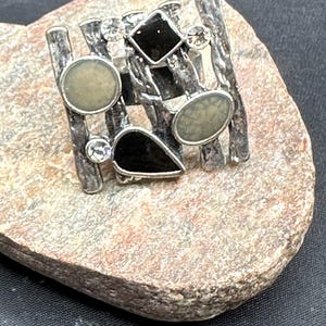 May include: Silver statement ring with a geometric design, black and cream accents, and clear crystals. The ring has a textured band. The ring is displayed on a stone surface.