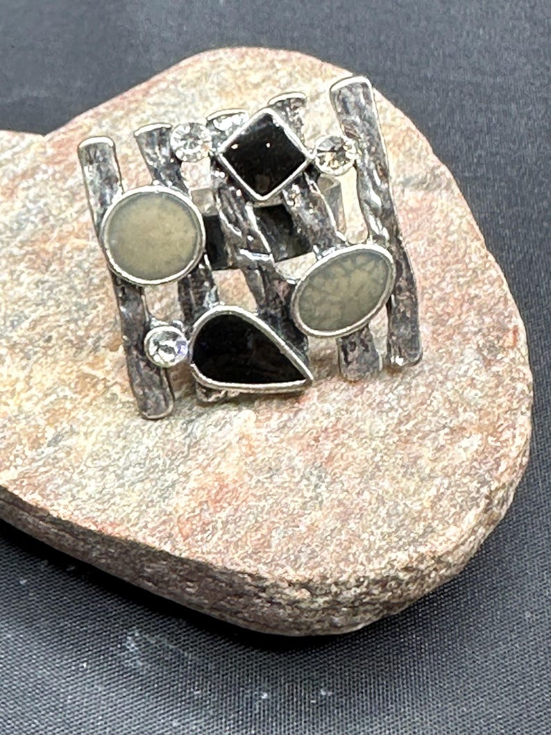 May include: Silver statement ring with a geometric design, black and cream accents, and clear crystals. The ring has a textured band. The ring is displayed on a stone surface.