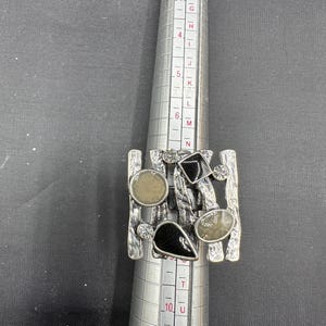 May include: A silver ring with an abstract design. It features a rectangular base with vertical bars and is adorned with black and grey stones. The ring is on a ring sizer. The ring sizer shows sizes from D to W.