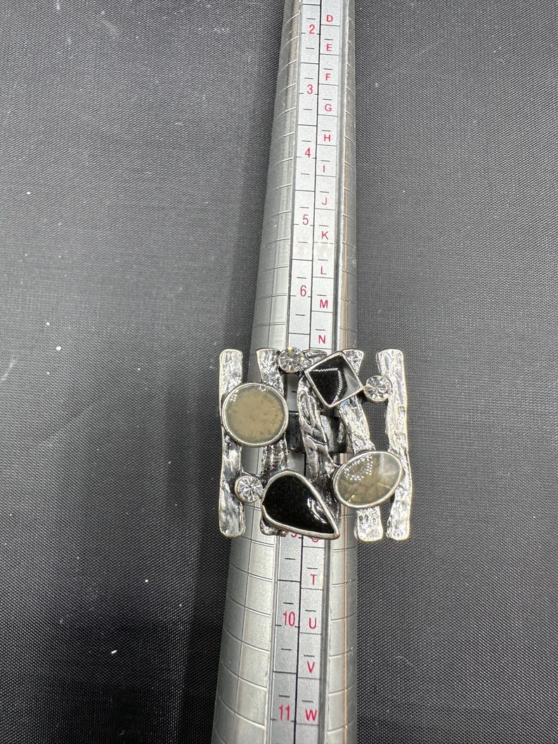 May include: A silver ring with an abstract design. It features a rectangular base with vertical bars and is adorned with black and grey stones. The ring is on a ring sizer. The ring sizer shows sizes from D to W.