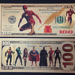 Peut inclure: Deux billets de 100 $ avec une finition en feuille d'or. Le billet du haut présente un super-héros rouge et jaune en costume rouge avec un éclair sur la poitrine. Le billet du bas présente un groupe de super-héros, dont Superman, Batman et Aquaman.