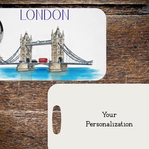 May include: Luggage tag featuring a watercolor illustration of the Tower Bridge in London, with the word "LONDON" in blue. A red double-decker bus and black car are on the bridge. The bottom tag is blank with the words "Your Personalization".
