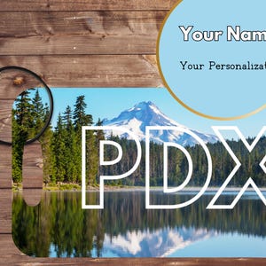 May include: A white key chain with a blue and green image of a mountain lake and a snowy mountain peak. The text "PDX" is printed in white on the image.