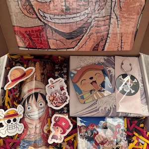 May include: An open cardboard box filled with One Piece merchandise. Includes a puzzle of Luffy's face, stickers, a small print, and a pin. The box is decorated with red and yellow shredded paper.