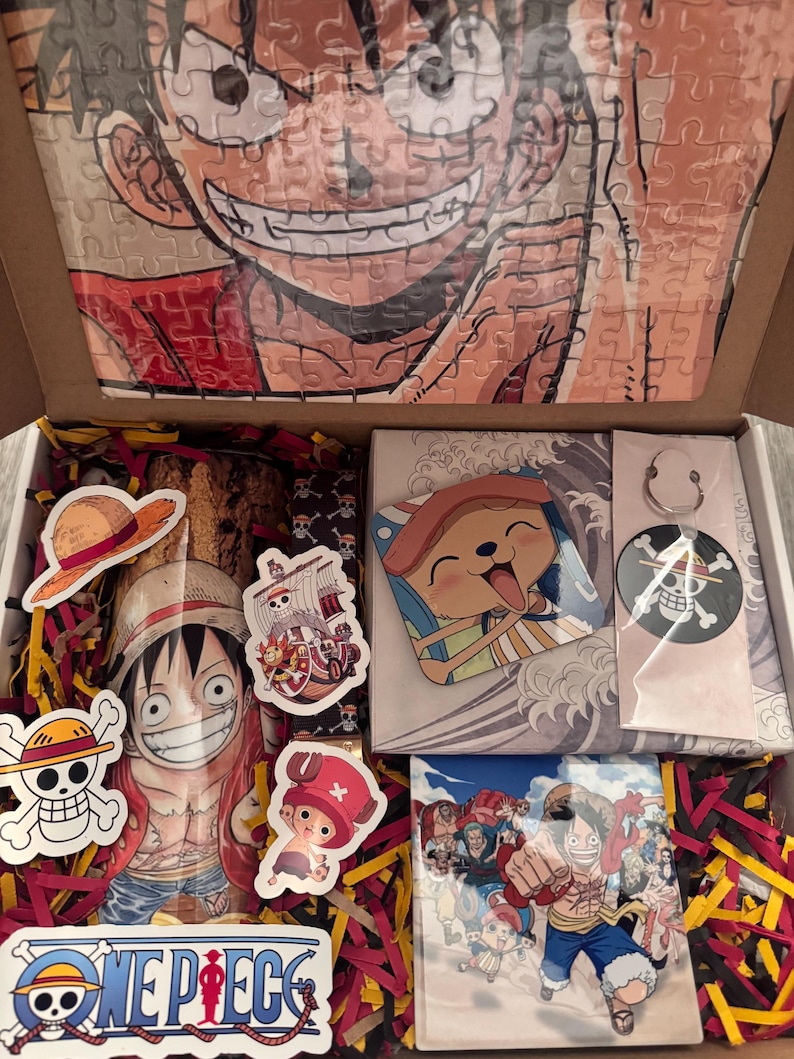May include: An open cardboard box filled with One Piece merchandise. Includes a puzzle of Luffy's face, stickers, a small print, and a pin. The box is decorated with red and yellow shredded paper.