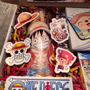 May include: An open box filled with red and yellow paper shreds, featuring One Piece themed stickers and a Luffy character print. Stickers include the Jolly Roger, a straw hat, and the ship, the Thousand Sunny. The box also contains a puzzle.