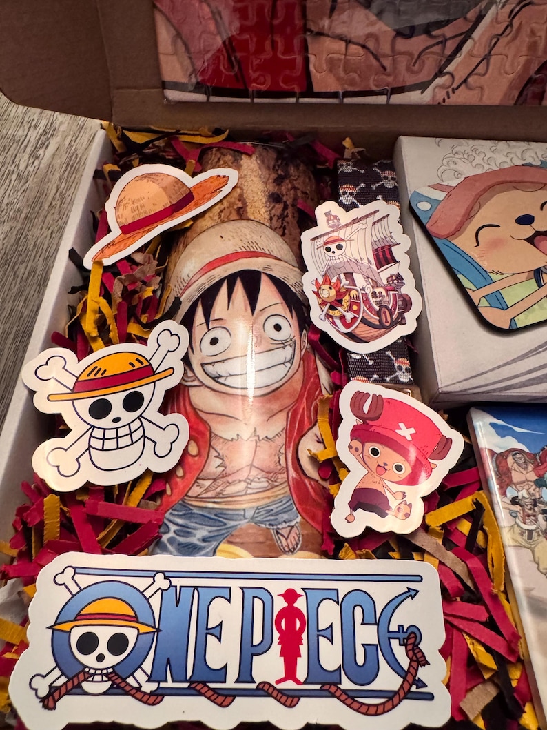 May include: An open box filled with red and yellow paper shreds, featuring One Piece themed stickers and a Luffy character print. Stickers include the Jolly Roger, a straw hat, and the ship, the Thousand Sunny. The box also contains a puzzle.