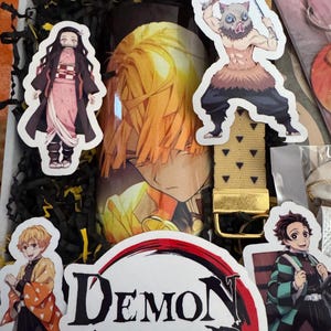 May include: A gift box with Demon Slayer merchandise. Includes a tumbler with a character's image, character stickers, and a gold-colored belt. The box is filled with black paper shreds.