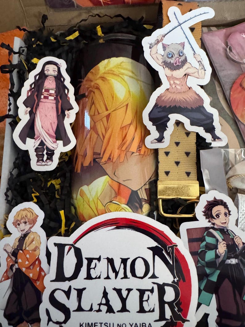May include: A gift box with Demon Slayer merchandise. Includes a tumbler with a character's image, character stickers, and a gold-colored belt. The box is filled with black paper shreds.