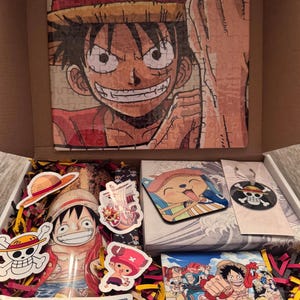 May include: An open cardboard box filled with One Piece merchandise. Includes a Luffy puzzle, stickers, a coaster, and a small print. The box is filled with red and yellow crinkle paper.
