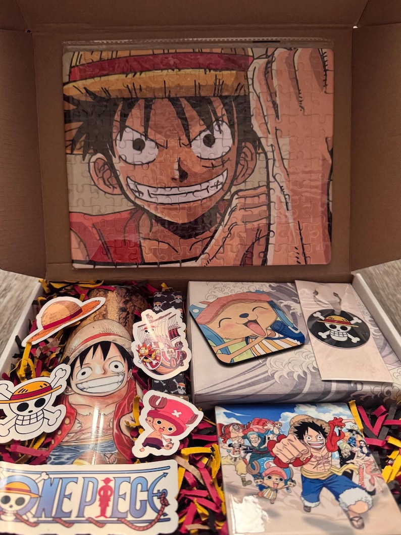 May include: An open cardboard box filled with One Piece merchandise. Includes a Luffy puzzle, stickers, a coaster, and a small print. The box is filled with red and yellow crinkle paper.