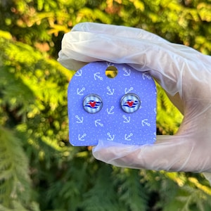 May include: A pair of stud earrings with a blue and red nautical themed design. The earrings are on a purple card with white anchor symbols.