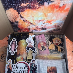 May include: An open box filled with Demon Slayer merchandise. Includes a puzzle with an anime character image, stickers, a keychain, and a tassel. The box also contains a bubble-wrapped package with a character image.
