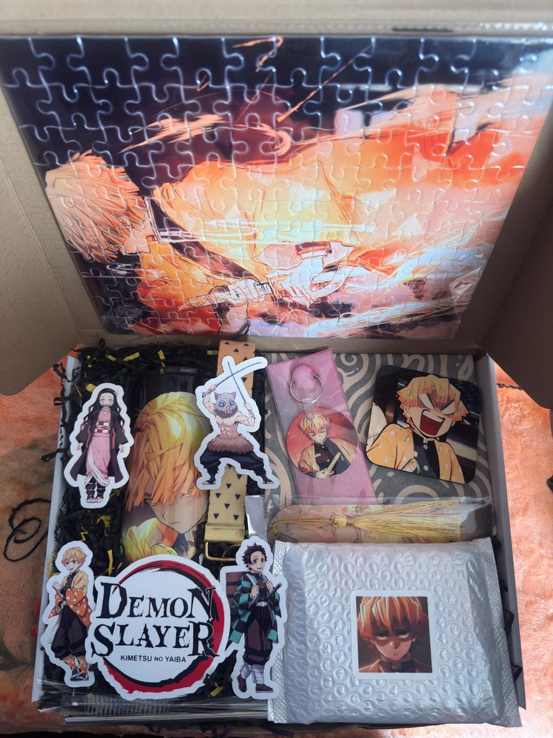 May include: An open box filled with Demon Slayer merchandise. Includes a puzzle with an anime character image, stickers, a keychain, and a tassel. The box also contains a bubble-wrapped package with a character image.