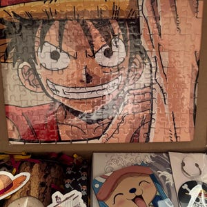 May include: A One Piece themed gift box featuring a puzzle of Luffy, stickers, a keychain, and a coaster. The puzzle shows Luffy smiling with his straw hat. The box contains various One Piece merchandise.