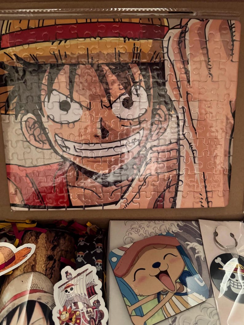 May include: A One Piece themed gift box featuring a puzzle of Luffy, stickers, a keychain, and a coaster. The puzzle shows Luffy smiling with his straw hat. The box contains various One Piece merchandise.