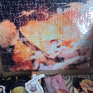 May include: A partially assembled jigsaw puzzle featuring a character with yellow hair and a sword, set against a fiery orange and yellow background. The puzzle is in a box with other character-themed merchandise.
