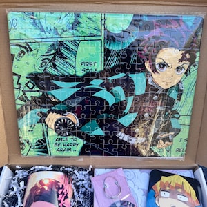 May include: A gift box set featuring a Demon Slayer anime puzzle, a mug with a character from the anime, a keychain with a round anime character charm, and a square coaster with a different anime character. The puzzle features a green and black background with a character holding a sword. The mug features a pink and white character with pink eyes. The keychain features a round charm with multiple anime characters. The coaster features a yellow and black character with a surprised expression.