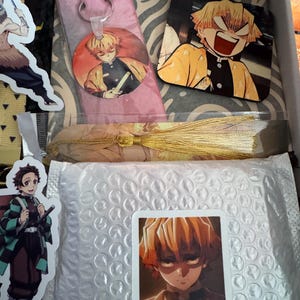 May include: A box filled with anime-themed merchandise. Includes a keychain, stickers, and a coaster featuring characters with yellow and orange hair. The items are packaged with bubble wrap and decorative paper.
