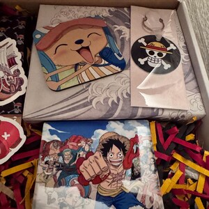 May include: An open box containing One Piece merchandise. Includes a Chopper character sticker, a keychain with a skull and crossbones logo, and a print featuring Luffy and other characters. The box is filled with colorful paper shreds.