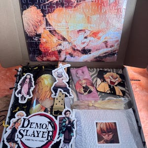 May include: An open box filled with Demon Slayer merchandise. Includes a puzzle with a character illustration, stickers, a keychain, and a bookmark. The box contains a variety of items featuring characters from the anime series.