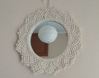 May include: A round mirror with a white macrame frame. The mirror is reflecting a white paper lantern.