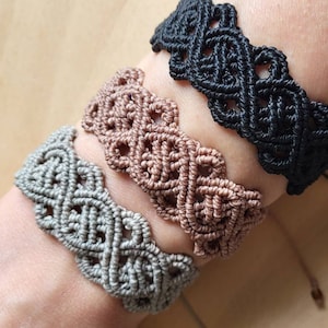 May include: Three macrame bracelets in different colors: gray, brown, and black. The bracelets are made with a knotting technique that creates a geometric pattern.