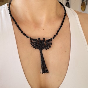 May include: A black macrame necklace with a bat pendant and a tassel. The bat is made of black cord and has a detailed design. The tassel is also made of black cord and hangs down from the bat.