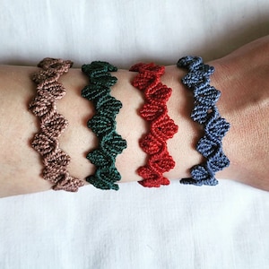 Könnte beinhalten: Vier handgefertigte Makramee-Armbänder in Braun-, Grün-, Rot- und Blautönen. Jedes Armband hat ein Zickzackmuster. Die Armbänder werden auf einem Arm vor weißem Hintergrund gezeigt.