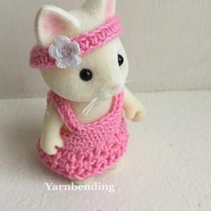 Puede incluir: Una figura de conejito blanco con un vestido y una diadema de ganchillo rosa con una flor blanca. El texto "Yarnbending" es visible en la parte inferior de la imagen. El conejito tiene ojos negros y orejas internas rosas.
