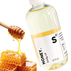 May include: A clear body oil bottle with a white label that reads "HONEY. body oil" and "10 fl oz". A wooden honey dipper drips honey onto a honeycomb.