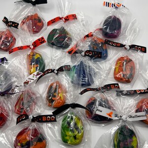 K&ouml;nnte beinhalten: Eine Sammlung kleiner, einzeln verpackter Halloween-Kreide. Jede Kreide ist in Form einer K&uuml;rbislaterne geformt und in verschiedenen Farben wie Orange, Gr&uuml;n, Lila und Schwarz gehalten. Die Kreide sind mit einem schwarz-wei&szlig;en Band gebunden und haben ein "BOO!"-Etikett.