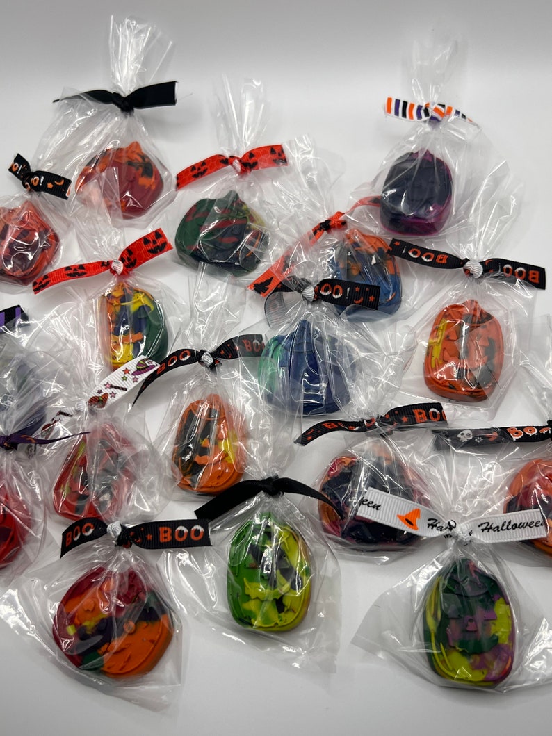 K&ouml;nnte beinhalten: Eine Sammlung kleiner, einzeln verpackter Halloween-Kreide. Jede Kreide ist in Form einer K&uuml;rbislaterne geformt und in verschiedenen Farben wie Orange, Gr&uuml;n, Lila und Schwarz gehalten. Die Kreide sind mit einem schwarz-wei&szlig;en Band gebunden und haben ein "BOO!"-Etikett.