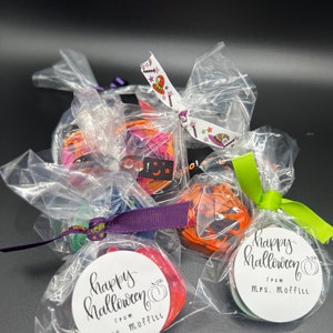 K&ouml;nnte beinhalten: Vier kleine durchsichtige Plastikt&uuml;ten gef&uuml;llt mit bunten Halloween-Leckereien, jede mit einem Band und einem Etikett gebunden, auf dem "Happy Halloween" mit einem K&uuml;rbis und "From Mrs. Moffitt" steht.