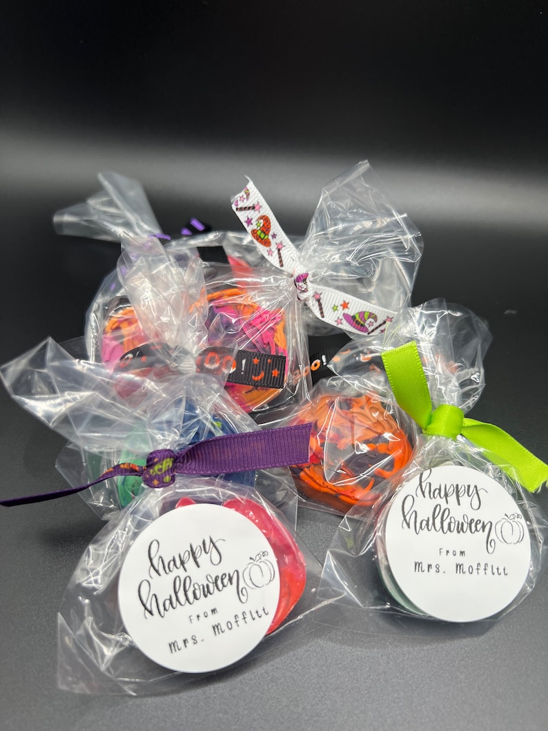 K&ouml;nnte beinhalten: Vier kleine durchsichtige Plastikt&uuml;ten gef&uuml;llt mit bunten Halloween-Leckereien, jede mit einem Band und einem Etikett gebunden, auf dem "Happy Halloween" mit einem K&uuml;rbis und "From Mrs. Moffitt" steht.