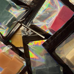 May include: An assortment of colorful, square-shaped wax melts in clear, iridescent packaging with black zippers. The melts are various colors, including green, pink, and beige. A white straw is visible in the center of the image.