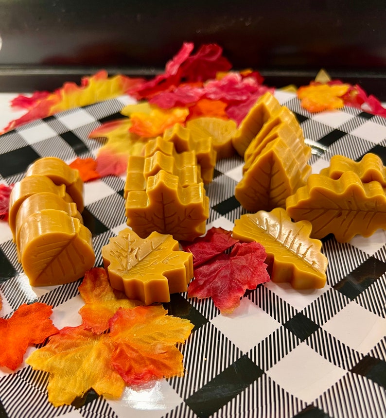 May include: A collection of golden yellow leaf-shaped wax melts, arranged on a black and white checkered surface with fall-colored leaves.