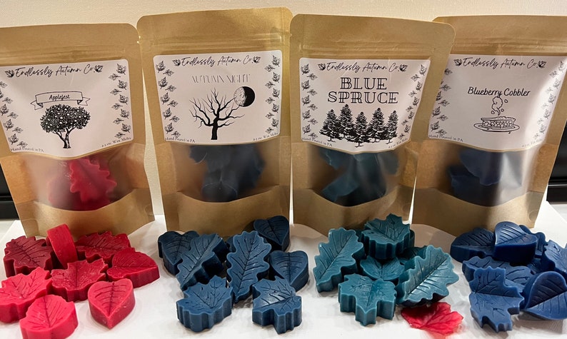May include: Four bags of wax melts in leaf shapes. The bags are brown paper with white labels. The labels have illustrations and text. The wax melts are red, blue, and teal. The scents are Applejest, Autumn Night, Blue Spruce, and Blueberry Cobbler.