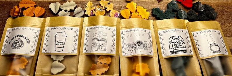 May include: Six brown paper bags with white labels and black illustrations. Each bag contains small wax melts shaped like leaves. The labels feature fall-themed designs and text, including "Pumpkin Pecan Waffle", "Pumpkin Spice Latte", "Sunflowers & Fall", "Cozy Winter", and "Tea Time".