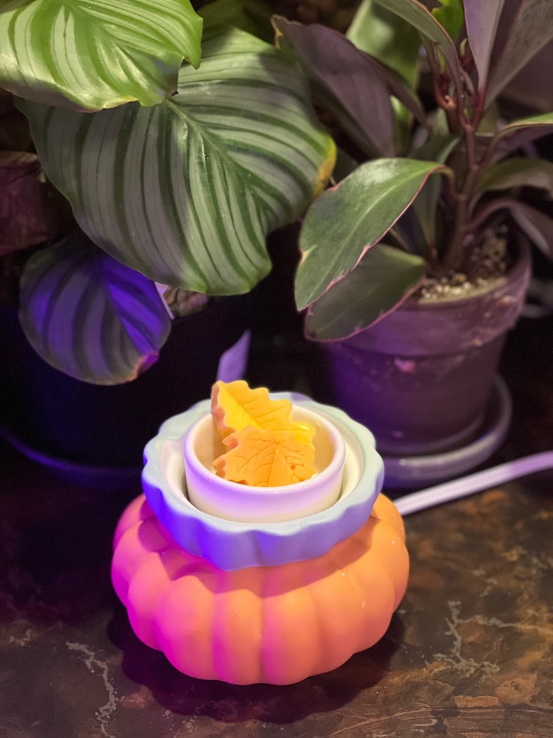 May include: A ceramic pumpkin-shaped wax warmer with a blue and white rim. The warmer has a small white dish inside with two yellow wax melts shaped like leaves.