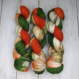 May include: Three skeins of yarn in green, orange, and white. The yarn is arranged in a row, with the green skein on the left, the orange skein in the middle, and the white skein on the right.