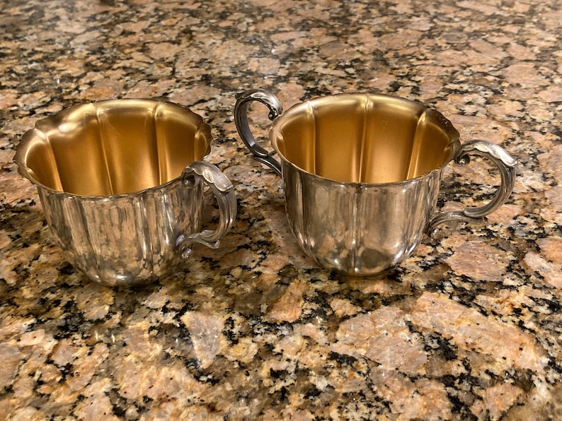 May include: Two silver sugar bowls with gold interiors. The bowls have a fluted design and ornate handles.