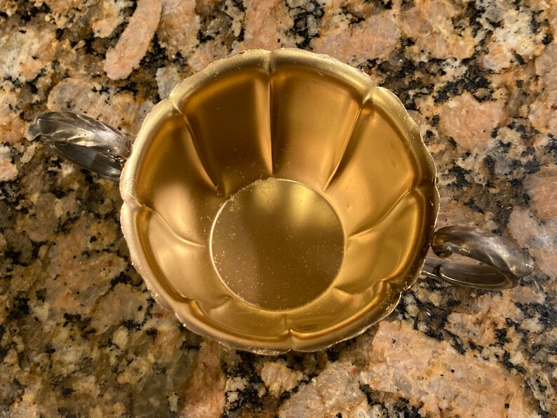 May include: A gold-colored metal sugar bowl with two handles. The bowl has a fluted design and a smooth, shiny finish.